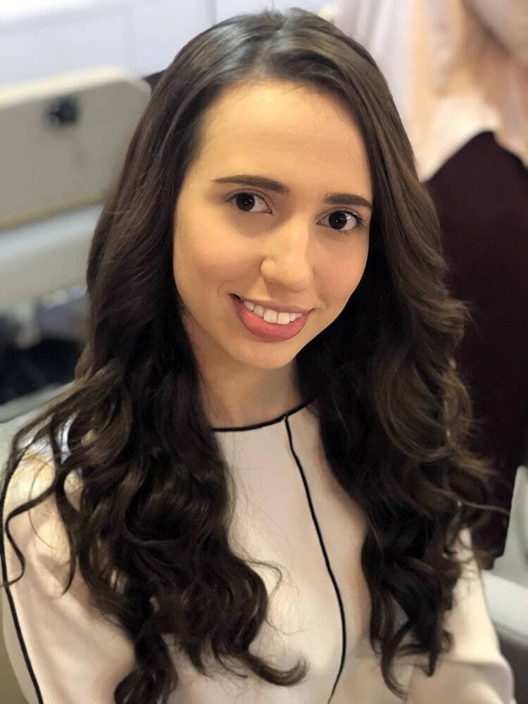 A blogueira Daniela Maksoud veste camisa branca com detalhes em costura preta e tem os cabelos castanhos estilizados com babyliss em ondas largas no estilo da modelo Gisele Bündchen.