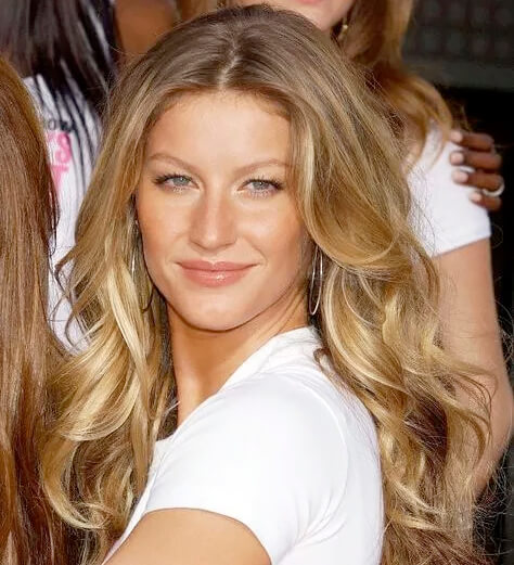 A modelo Gisele Bündchen usa brincos de argola prateada, veste uma camiseta branca e tem os cabelos loiros estilizados com babyliss em ondas largas.