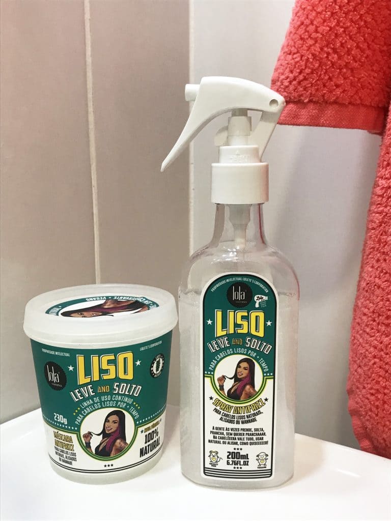 Máscara e spray antifrizz da linha Liso, Leve and Solto da Lola Cosmetics em cima da pia de um banheiro com uma toalha ao fundo.