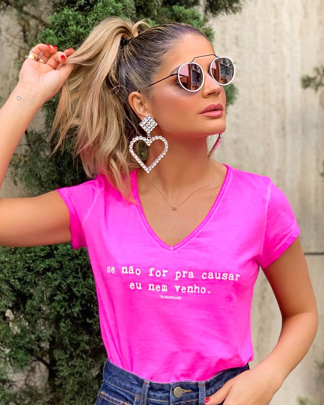 Thássia Naves usa cabelo preso em rabo de cavalo alto, camiseta rosa pink escrito se não for pra causar, eu nem venho, maxi brincos de coração com strass prata e óculos de sol.