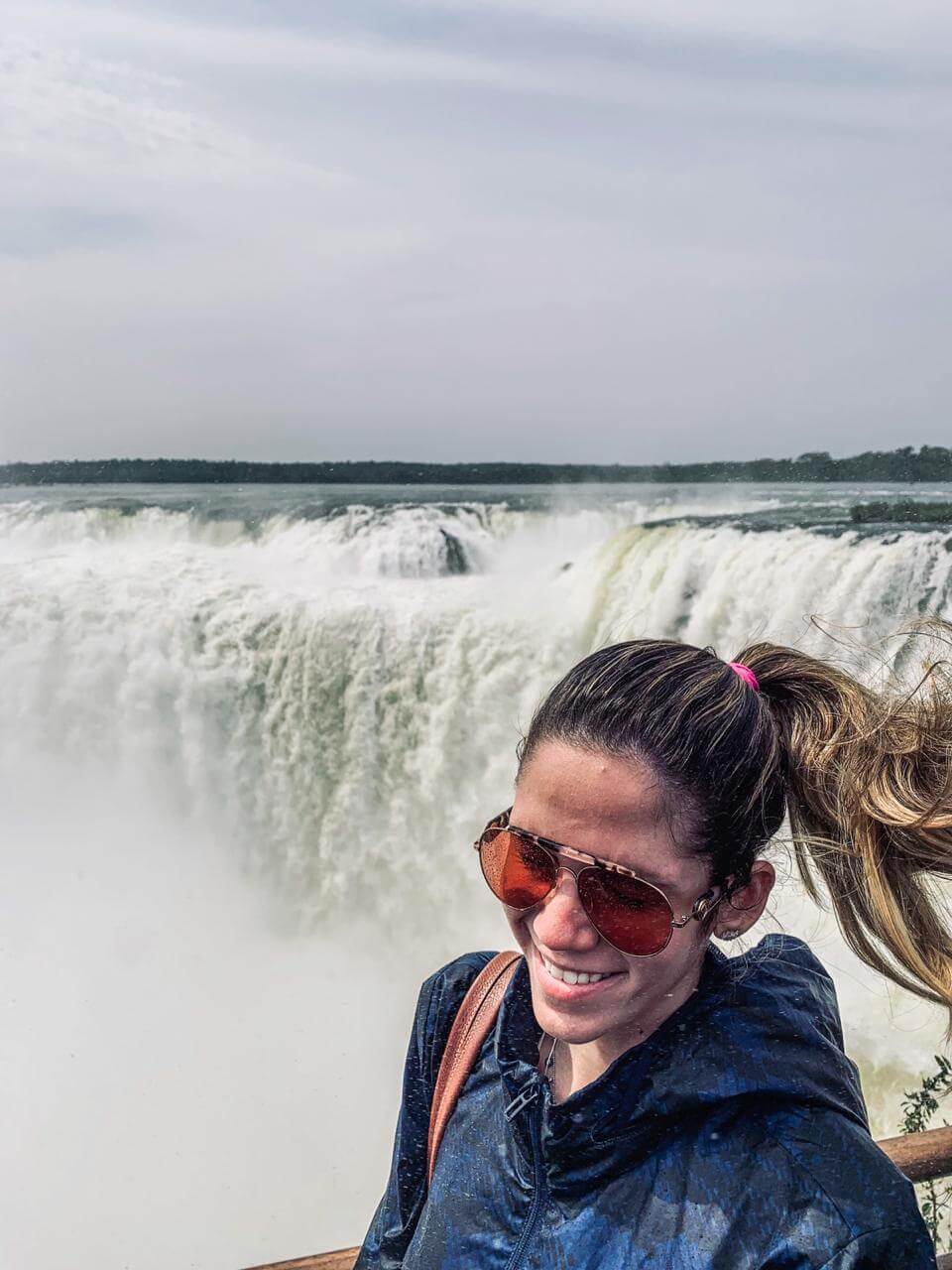 Manuela Brasão posa para foto sorridente com rabo de cavalo ao vento e cataratas de Foz do Iguaçu ao fundo.